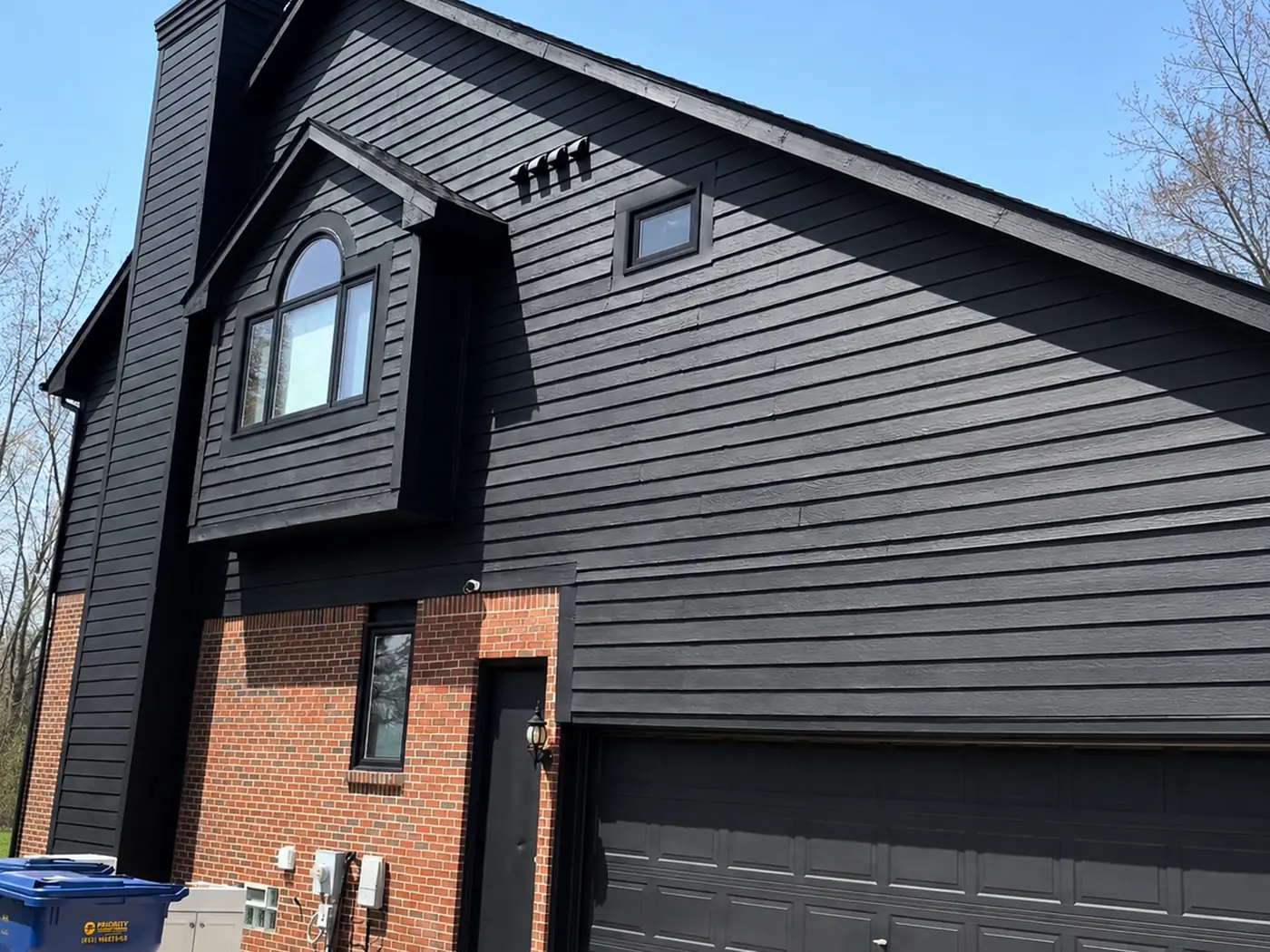 The same elevation after painting: siding, gable, dormer surround, and garage door all in a matte black that leaves the red brick as the only colour.